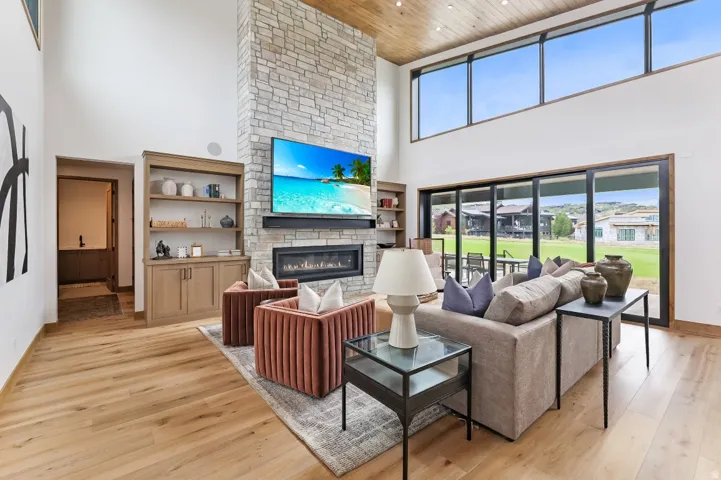 Vaulted ceiling with wood-finish, stone-clad fireplace with linear hearth, expansive floor-to-ceiling windows, built-in shelving, and wood-finish flooring