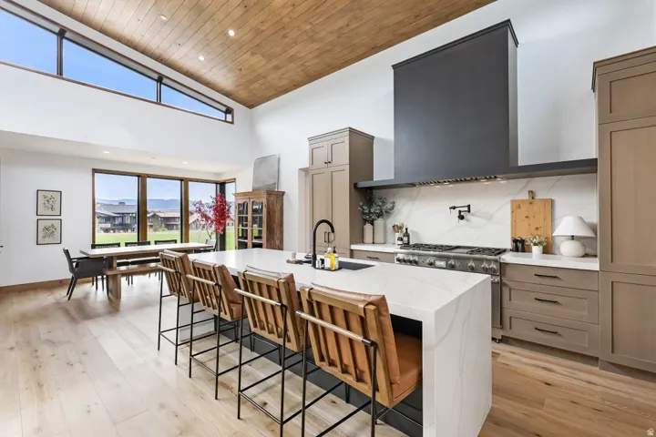 Vaulted wood-paneled ceiling, expansive window wall, light wood-finish flooring, large kitchen island with countertop, and professional-grade range with a substantial range hood