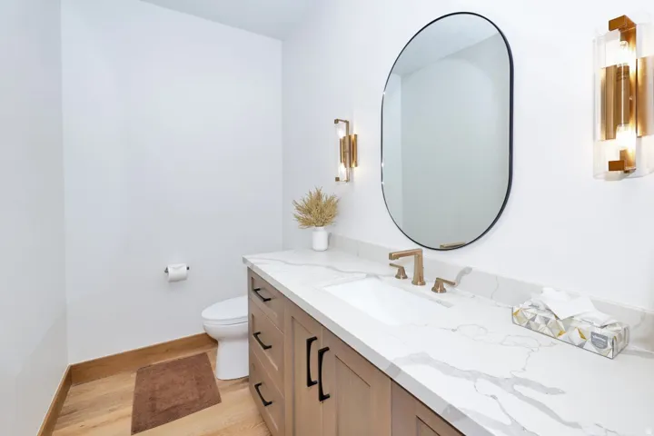 Wood-finish flooring, floating vanity with black hardware, integrated sink, quartz countertop, and a contemporary oval mirror with sconce lighting