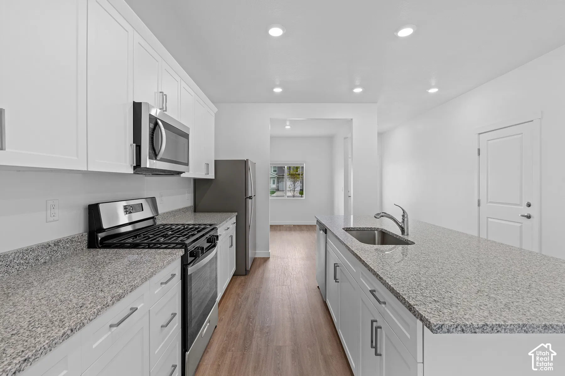 White shaker cabinetry with stainless steel hardware, stainless steel appliances, speckled stone countertops, a kitchen island with an undermount sink, and wood-finish flooring