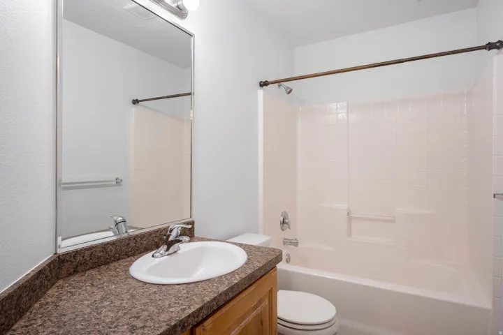 Bathroom featuring a white ceramic drop-in sink, chrome faucet, brown speckled countertop, wood-finish vanity, and a tub/shower combination with white tiled walls