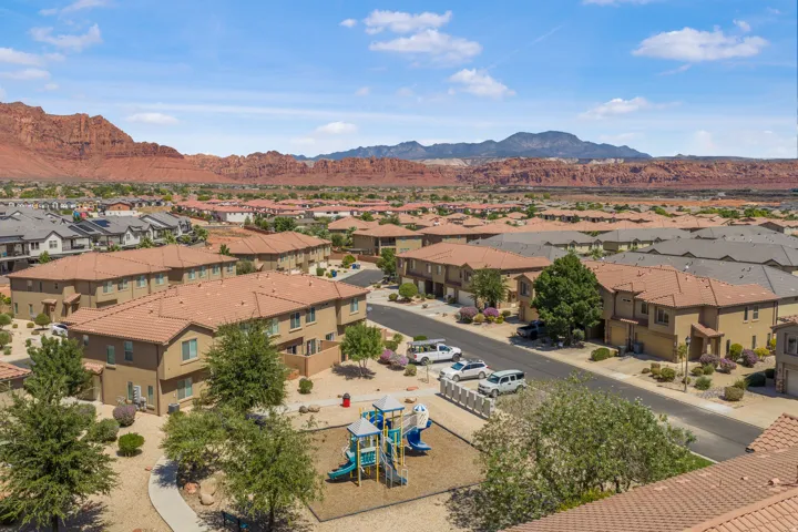 Community playground with play structure, surrounding residential structures with tiled roofs, and mature landscaping, all set against a backdrop of distant red rock formations