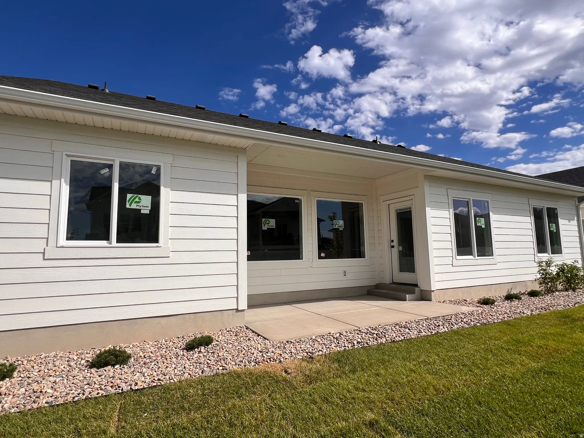 Exterior showcasing white siding, a concrete patio, multiple windows, and a landscaped rock bed with plantings