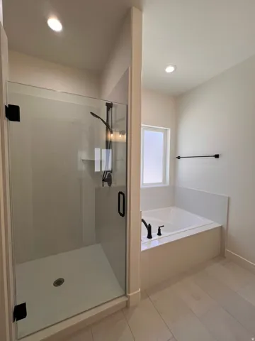 Walk-in shower with clear glass enclosure and matte black hardware, built-in soaking tub with matte black fixtures, and light-toned tile flooring