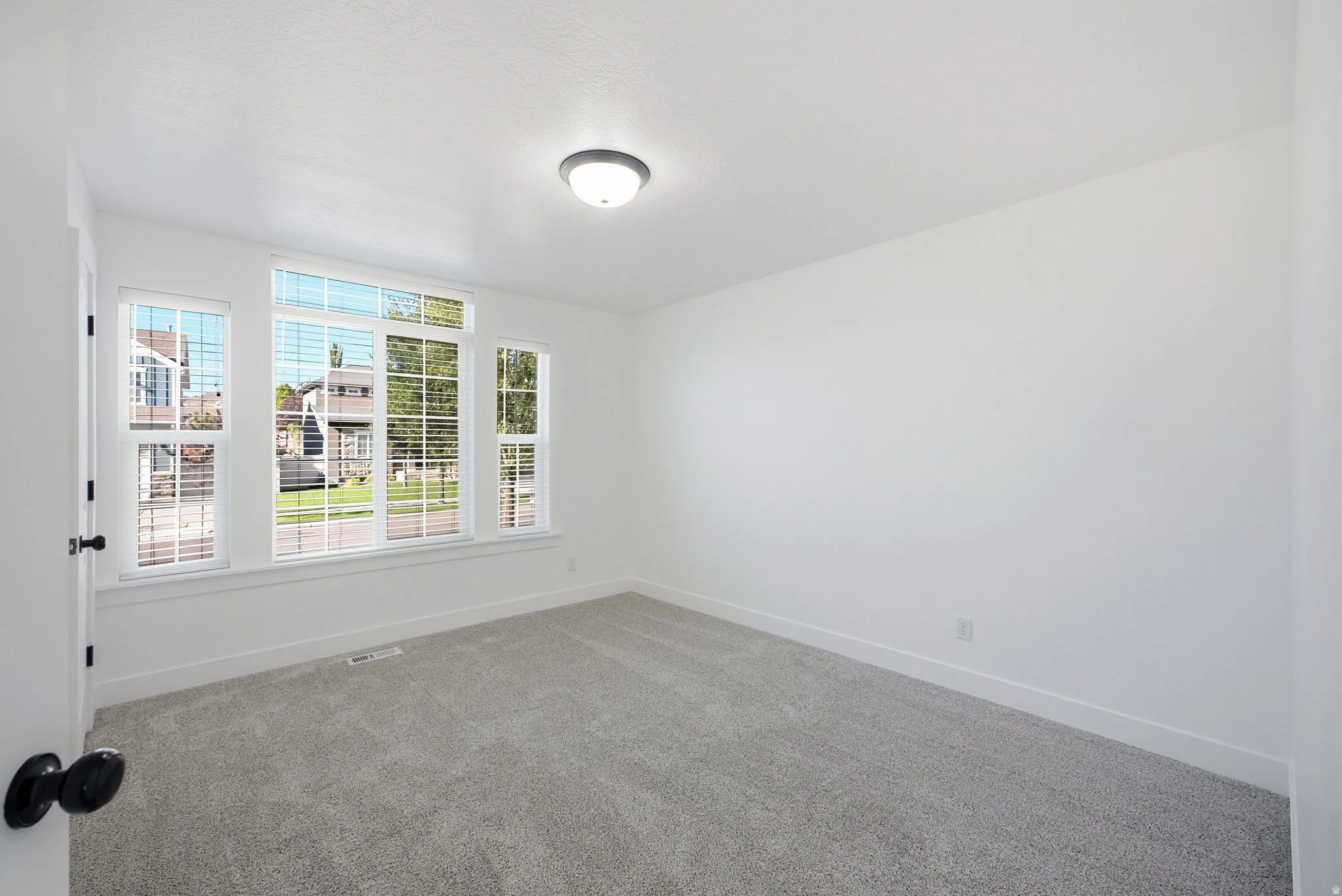 Recessed lighting, large window with blinds, light gray carpeting, white walls, and a white door with black hardware