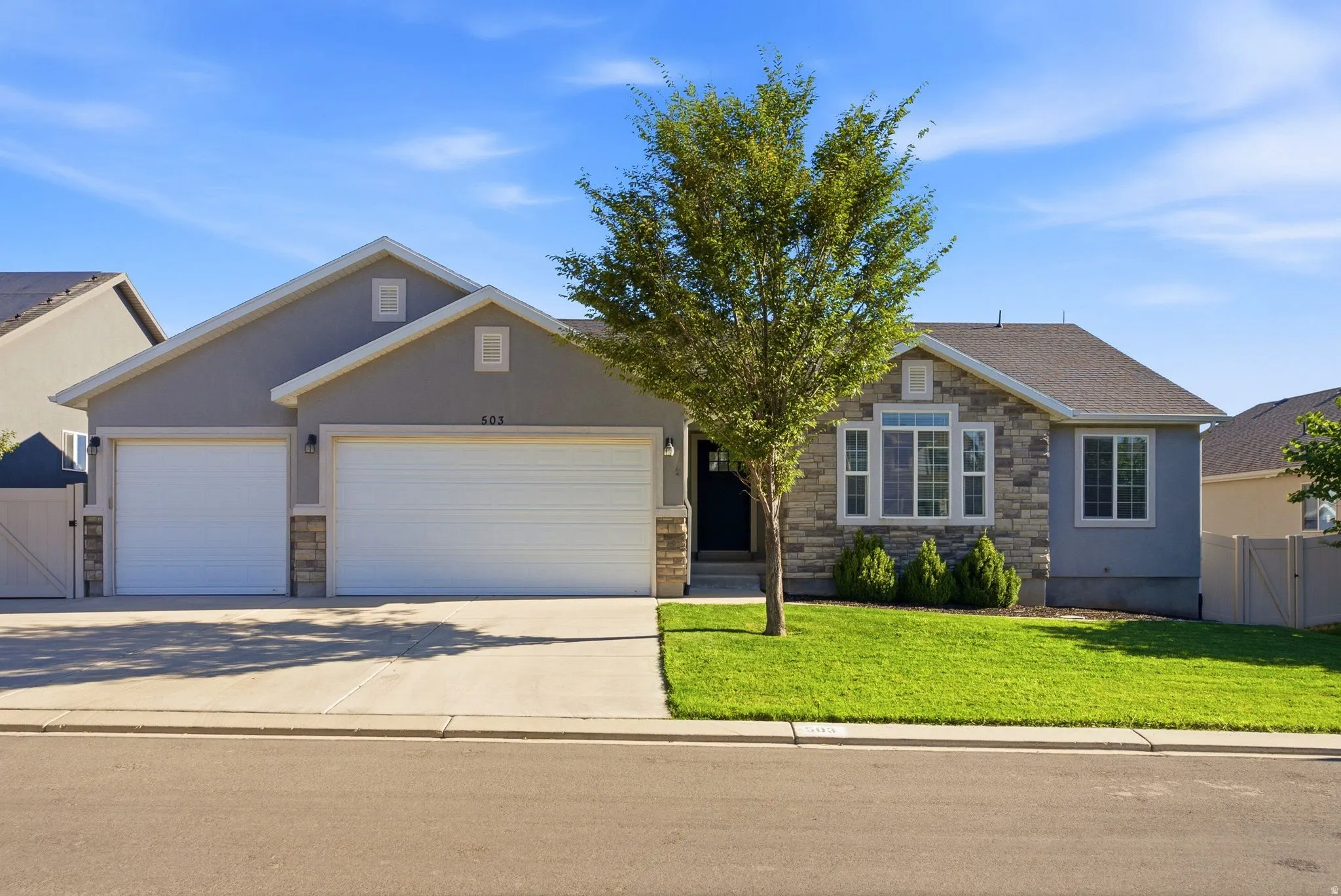 Two-car garage with white doors, stone façade accents, bay window, landscaped front yard, and privacy fencing