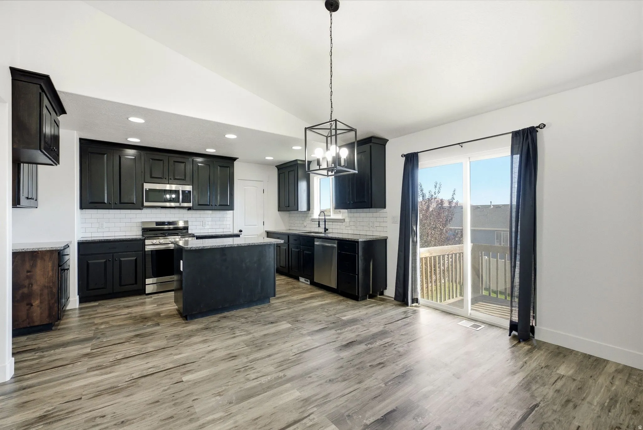 Vaulted ceiling kitchen featuring dark cabinetry, stainless steel appliances, a center island with countertop, wood-finish flooring, and a sliding glass door leading to a deck