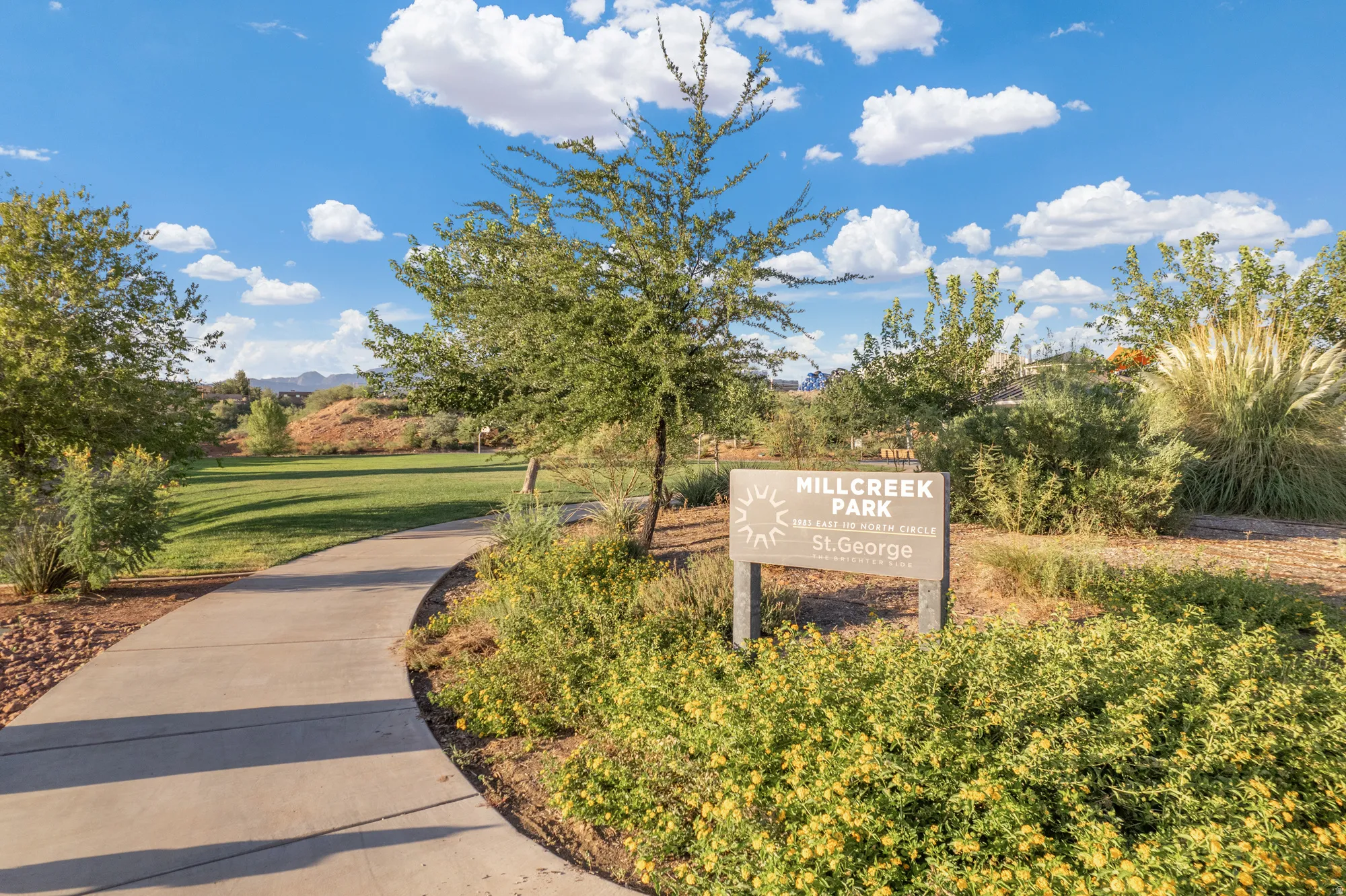 Paved walking path bordered by lush landscaping, leading to an expansive grassy park area
