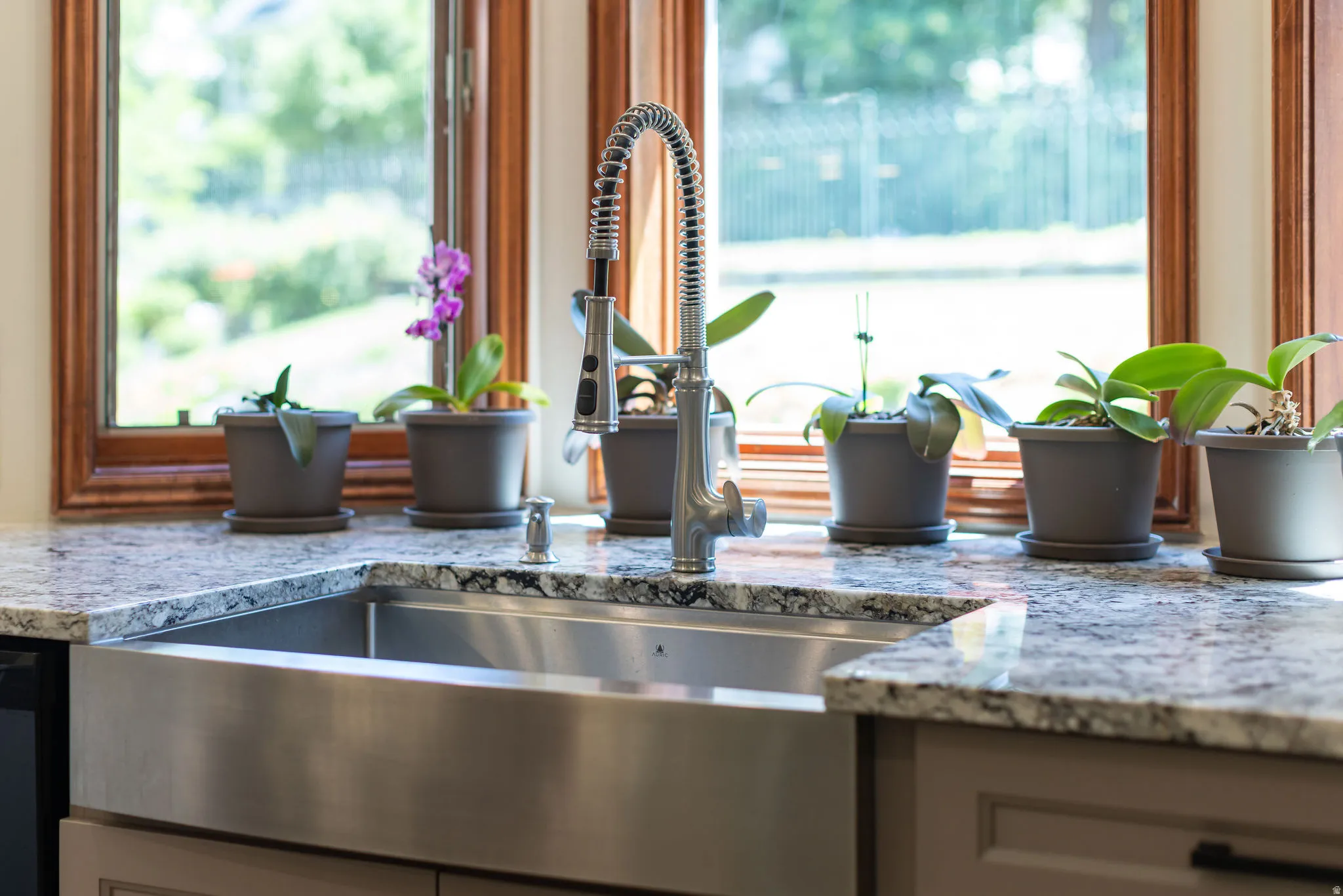 Farmhouse-style stainless steel sink with a high-arc pull-down faucet, set within variegated stone countertops, with wood-framed windows