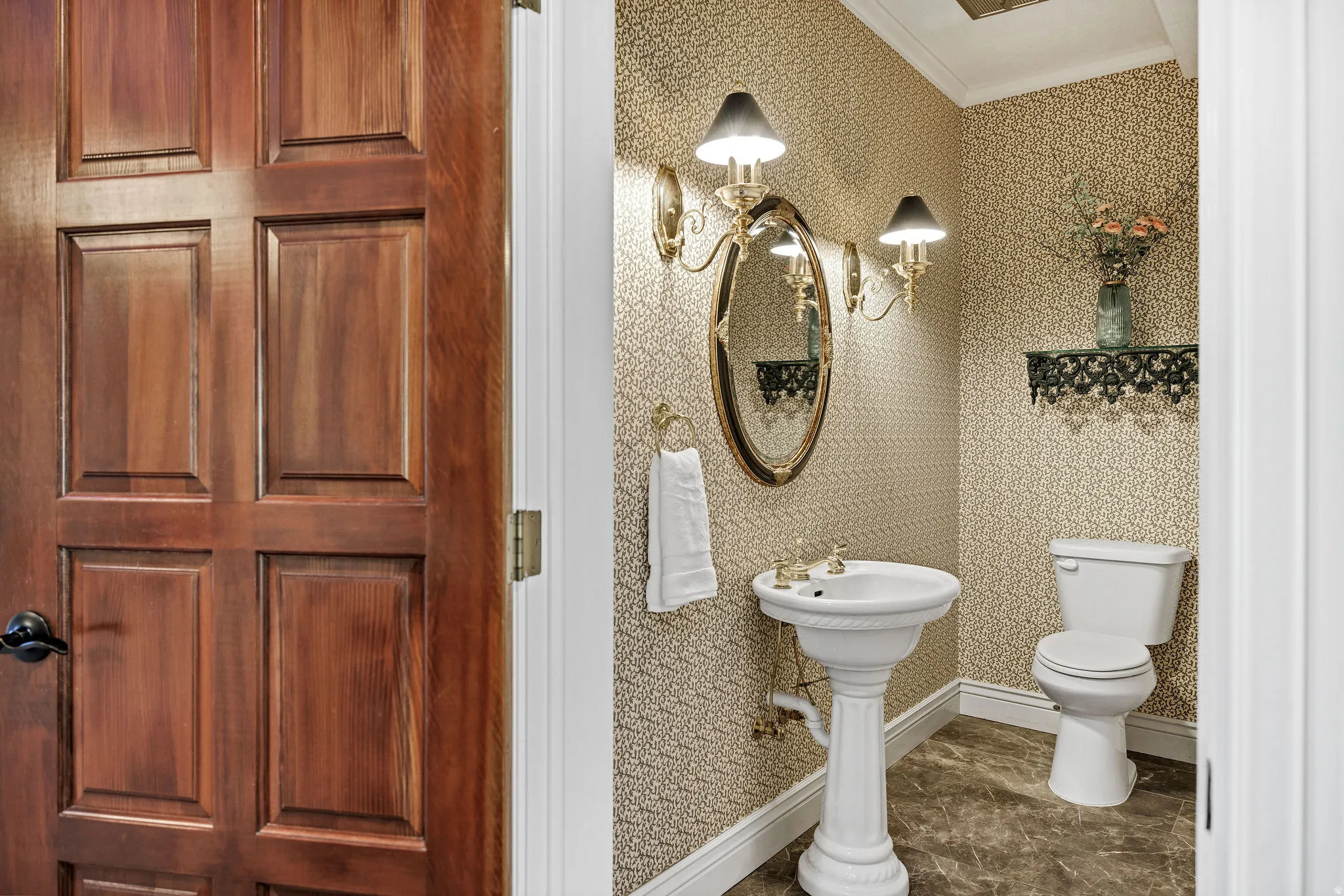 Paneled wood door opening to an interior featuring patterned wallpaper, pedestal sink, and wall-mounted lighting fixtures