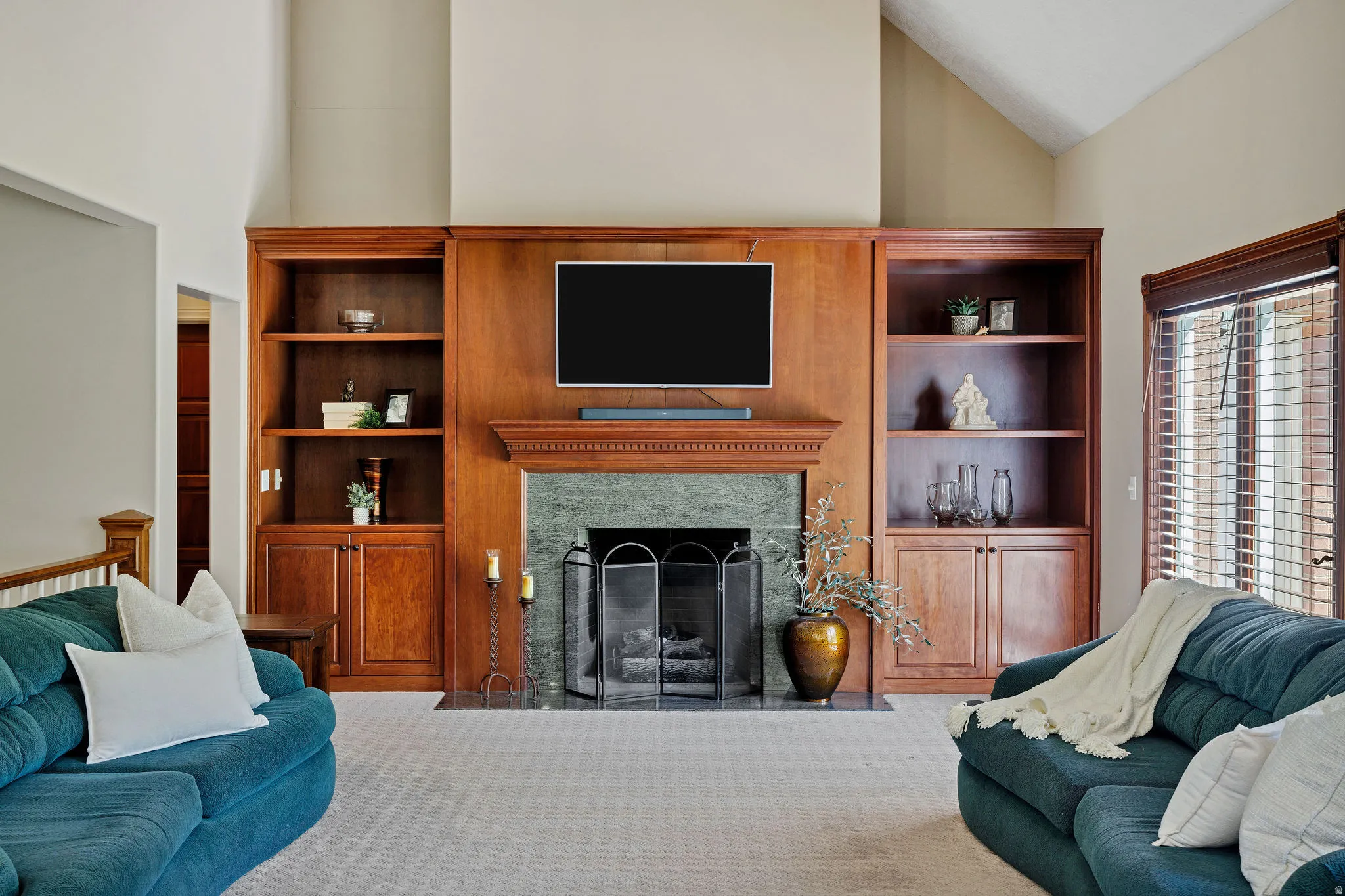 Vaulted ceiling, built-in shelving, stone fireplace surround, wood mantel, and window with blinds
