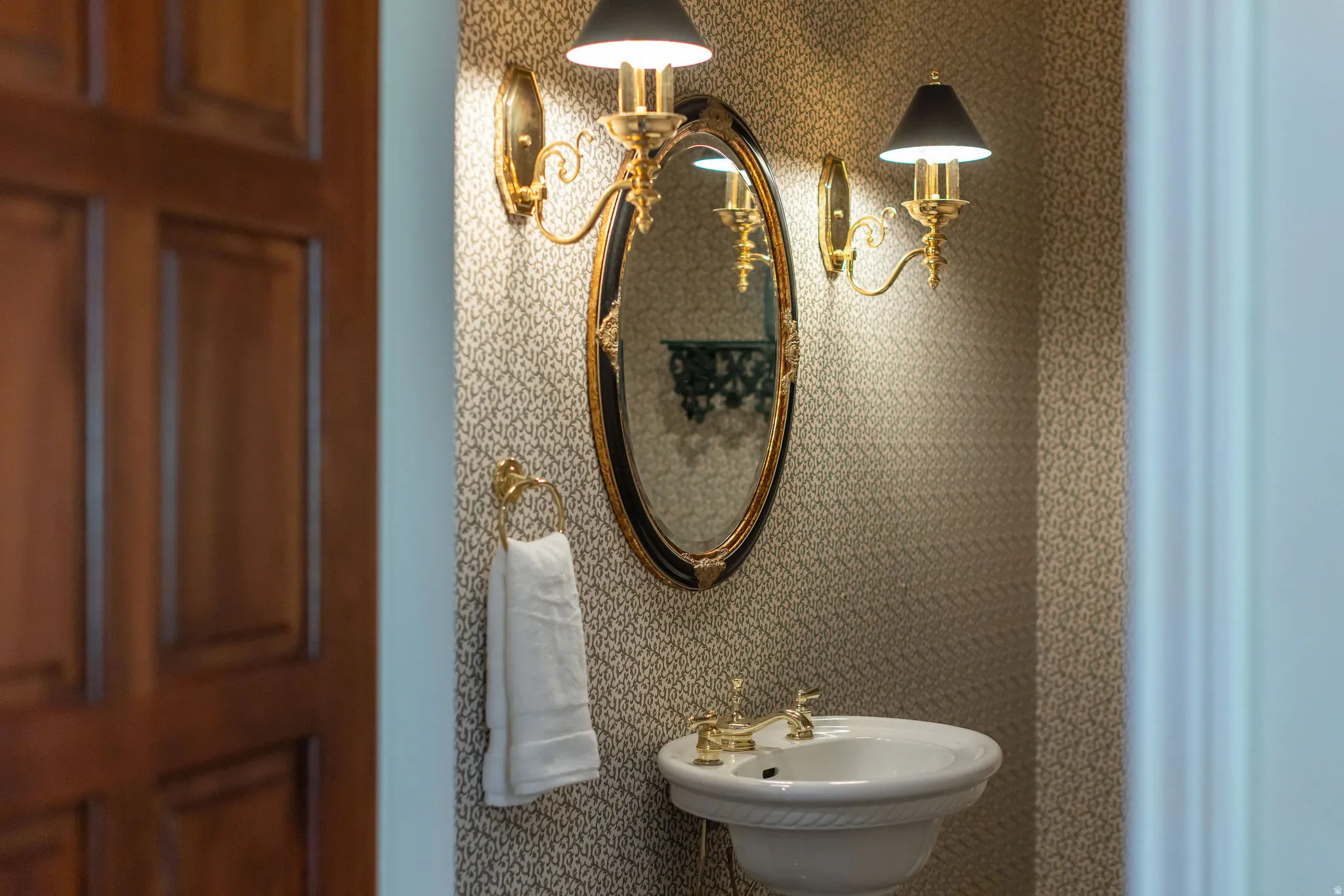Pedestal sink with brass-finish hardware, oval framed mirror, and two wall sconces with black shades
