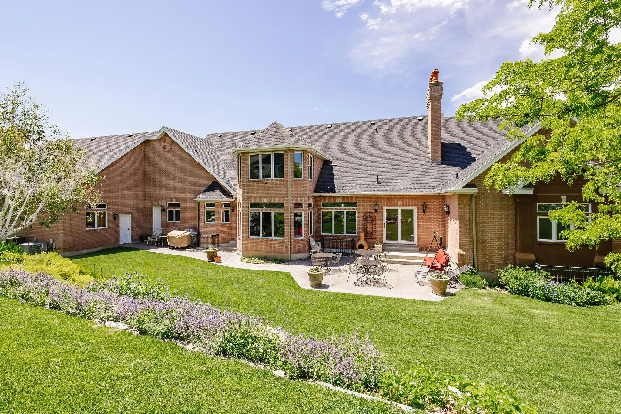 Expansive rear facade featuring brick construction, a multi-level roofline, a tiered concrete patio, and a landscaped lawn