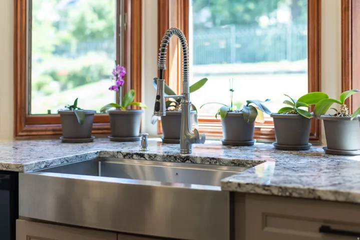 Farmhouse-style stainless steel sink with a high-arc pull-down faucet, set within variegated stone countertops, with wood-framed windows