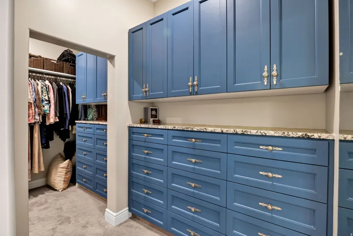 Luxurious walk-in closet featuring custom blue cabinetry with brushed gold hardware, abundant drawer storage, granite countertop, and neutral tile flooring