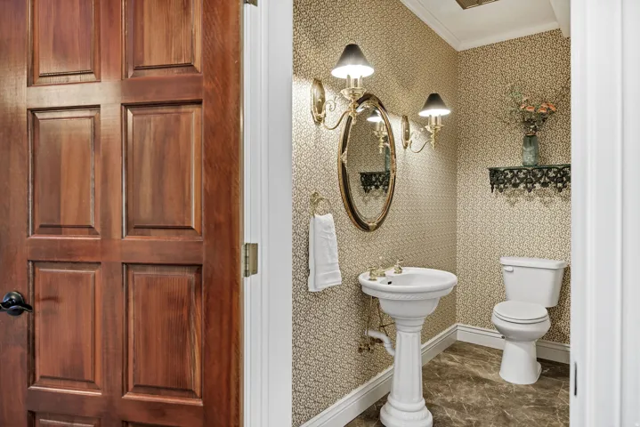 Paneled wood door opening to an interior featuring patterned wallpaper, pedestal sink, and wall-mounted lighting fixtures