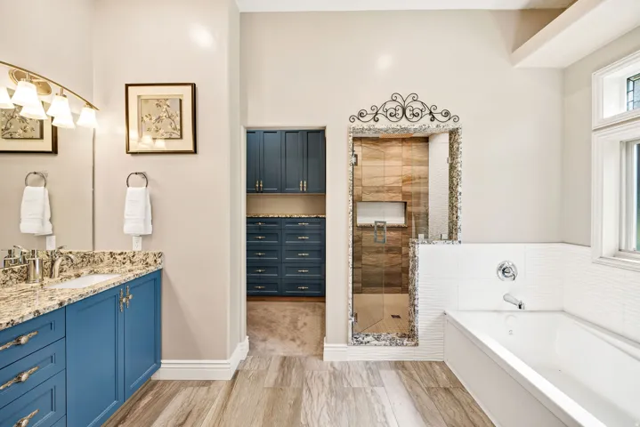Dual vanity with granite countertop, wood-finish flooring, built-in soaking tub, walk-in shower with tile surround, and custom cabinetry in an adjacent space
