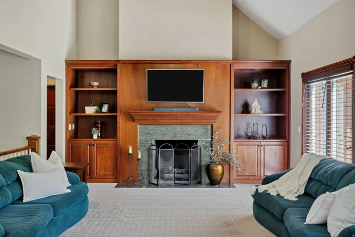 Vaulted ceiling, built-in shelving, stone fireplace surround, wood mantel, and window with blinds