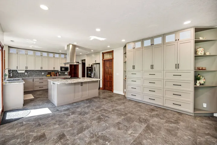 Spacious kitchen featuring an expansive central island, extensive light-toned cabinetry with dark hardware, a tile backsplash, and dark-toned stone-finish flooring