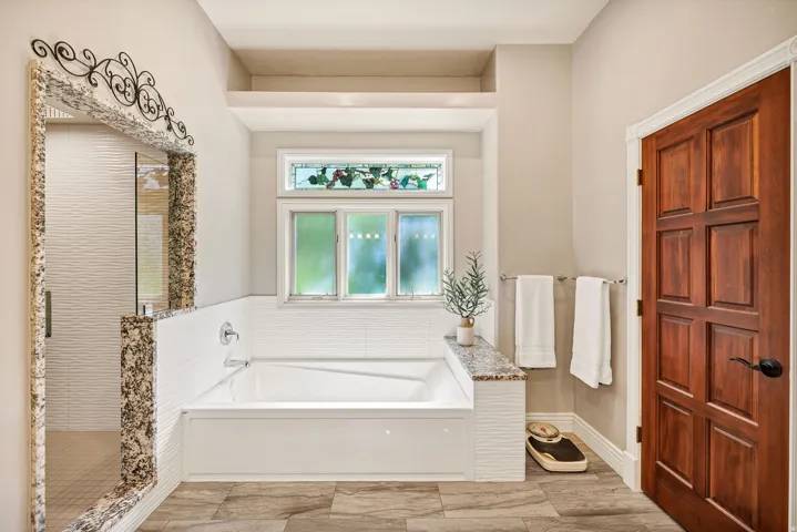 Soaking tub with a granite surround, integrated tiled shower, wood-finish flooring, and a decorative stained-glass window