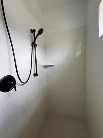Walk-in shower with textured tile walls, matte black shower fixtures, a handheld showerhead, and an integrated corner shelf