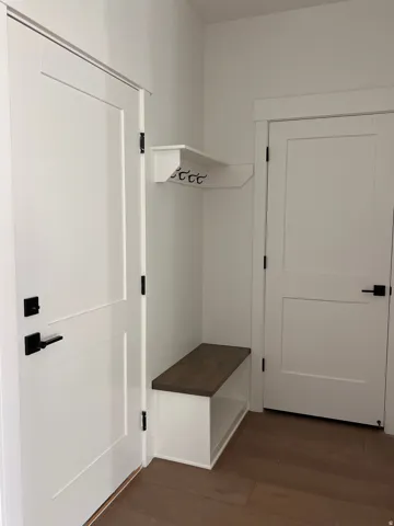 Dedicated entryway featuring a built-in bench with wood-finish seating, overhead wall-mounted hooks for storage, white painted doors with black hardware, and wood-finish flooring throughout