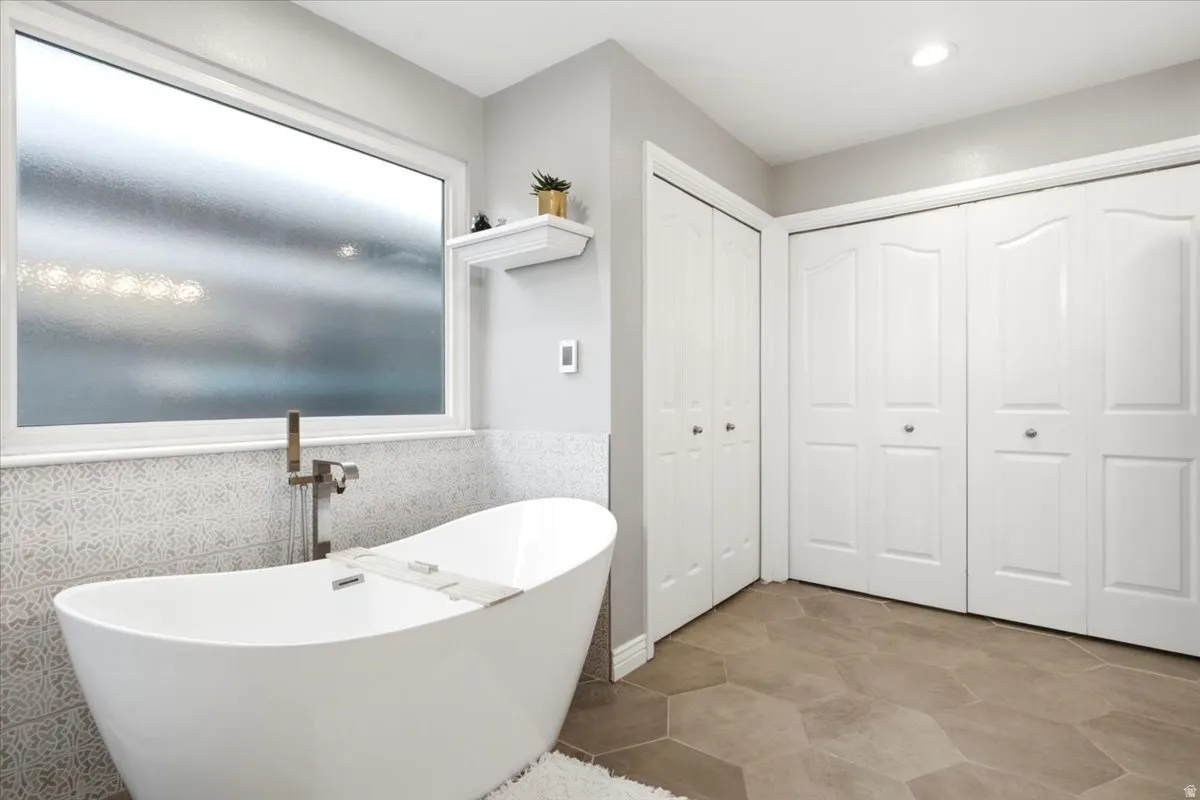 Freestanding soaking tub with floor-mounted faucet, privacy window, decorative tile wainscoting, and heated tile flooring