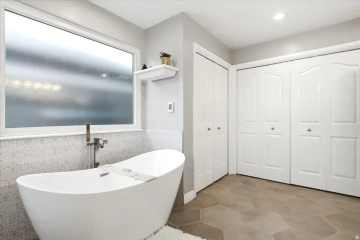 Freestanding soaking tub with floor-mounted faucet, privacy window, decorative tile wainscoting, and heated tile flooring