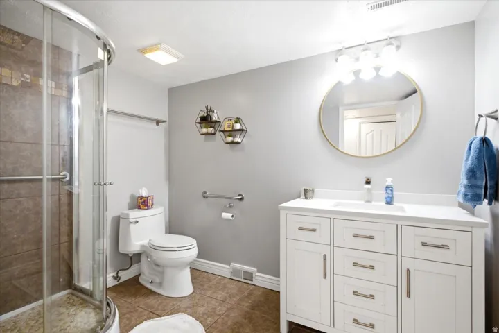Curved glass shower enclosure, white vanity with integrated sink, gold-framed circular mirror, three-light vanity fixture, and tile flooring