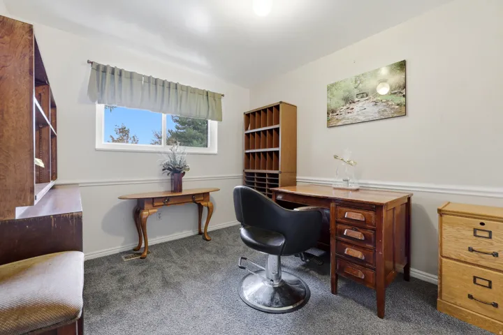 Versatile home office featuring dual wood-finish desks, built-in shelving with display storage, natural light from large windows, and carpeted flooring