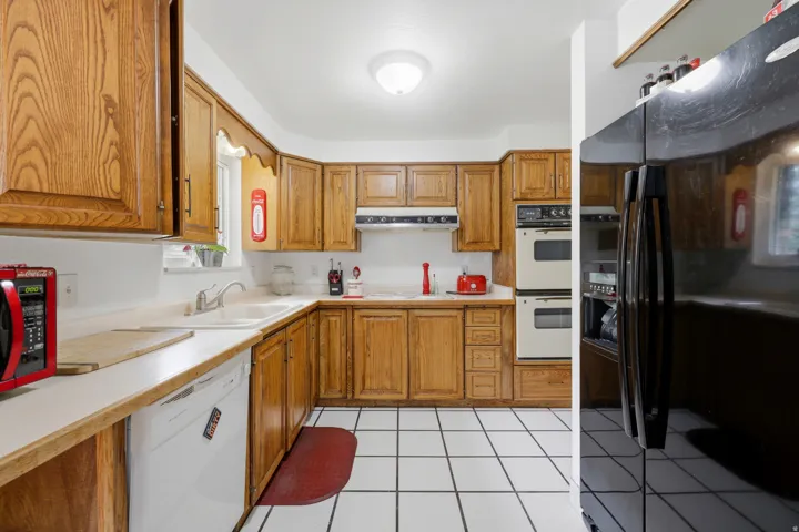 Galley-style kitchen featuring wood cabinetry, white tile flooring, stainless steel appliances including double wall ovens, and a ceramic sink with under-counter storage