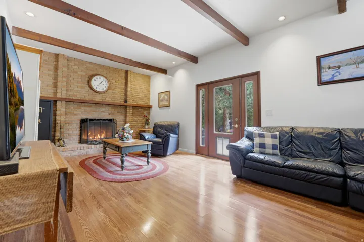 Spacious living room featuring exposed wooden beam ceiling details, hardwood flooring throughout, a brick fireplace with mantel, recessed lighting, and glass French doors providing natural light and access to outdoor spaces