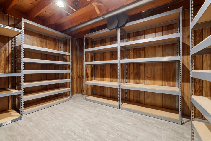 Spacious storage room featuring industrial-grade metal shelving with white finish throughout three walls, concrete flooring, exposed wood-beam ceiling with recessed lighting, and ample square footage for organization and inventory management