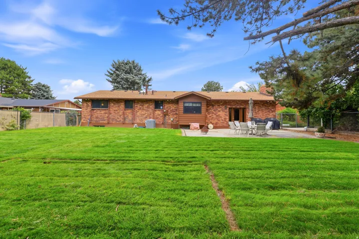 Expansive manicured lawn with mature landscaping and tree coverage, complemented by a single-story brick residence featuring multiple windows and a covered patio area with outdoor seating space