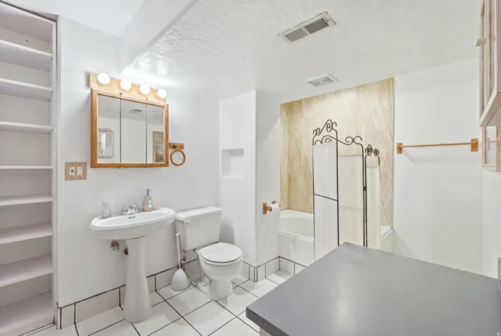 Pedestal sink with chrome faucet, framed medicine cabinet with vanity lighting, built-in shelving, white tiled flooring, and a tub with tiled surround