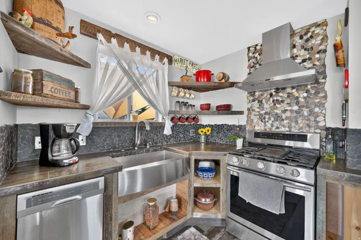 Stainless steel apron front sink, pebble tile backsplash, wood-finish open shelving, stainless steel dishwasher, and stainless steel gas range with exhaust hood