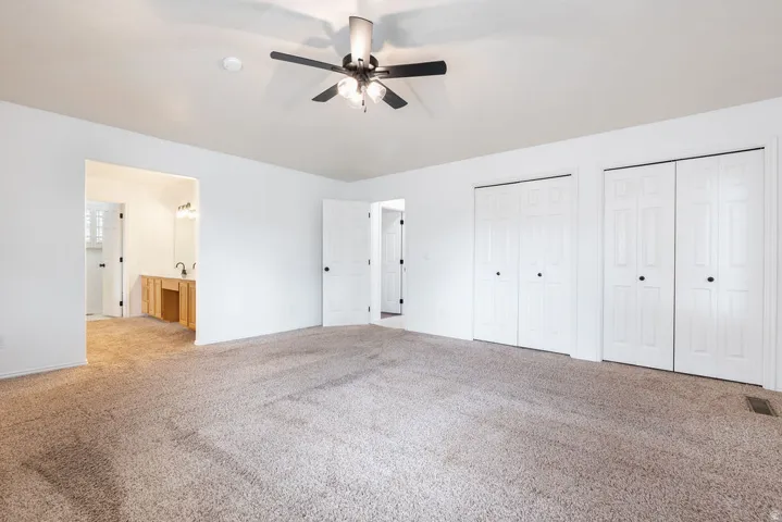 Expansive carpeted area featuring a ceiling fan, two sets of bi-fold closet doors, and an open doorway to a bathroom with a wood-finish vanity and dual sinks