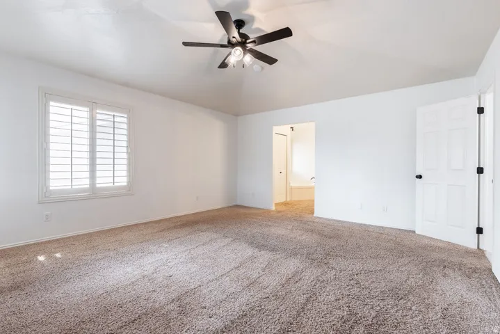 Spacious bedroom showcasing neutral wall finishes, plush carpeting, ceiling fan with integrated lighting, shuttered window with natural light, and direct access to adjoining spaces through multiple doorways