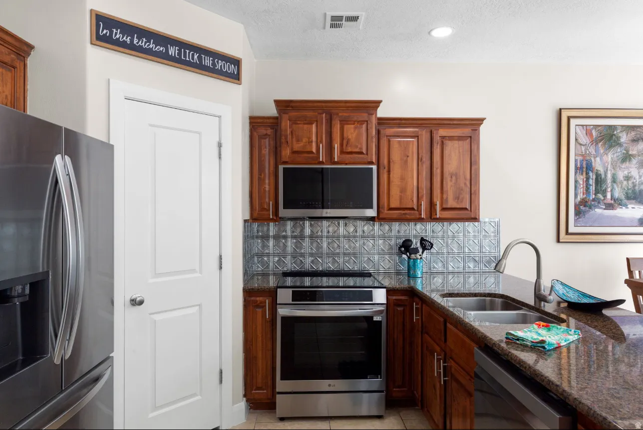 Stainless steel appliances, rich wood cabinetry, patterned metallic backsplash, granite-style countertops, and a stainless steel sink with a high-arch faucet