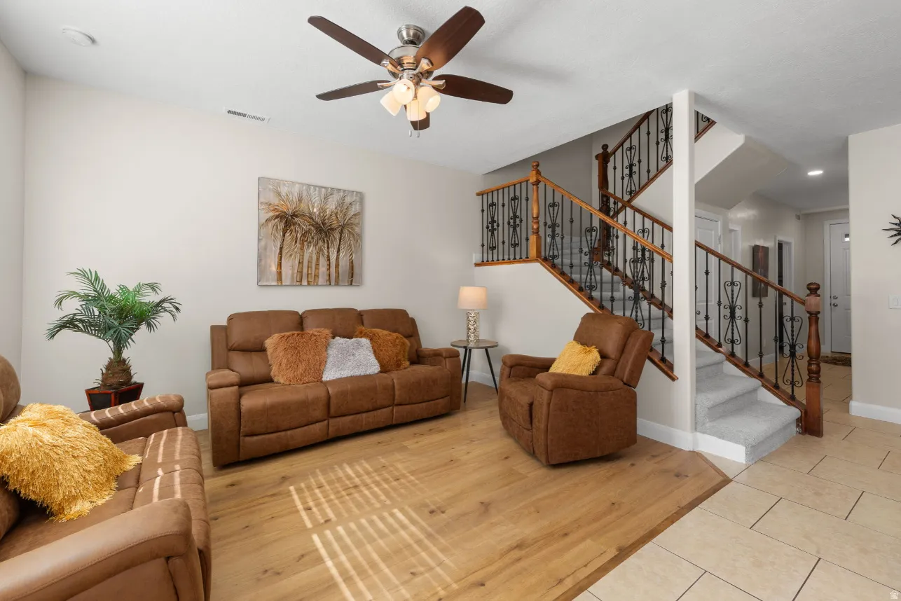 Spacious living area featuring wood-finish flooring, a ceiling fan with integrated lighting, and a staircase with decorative wrought iron spindles and wood handrails