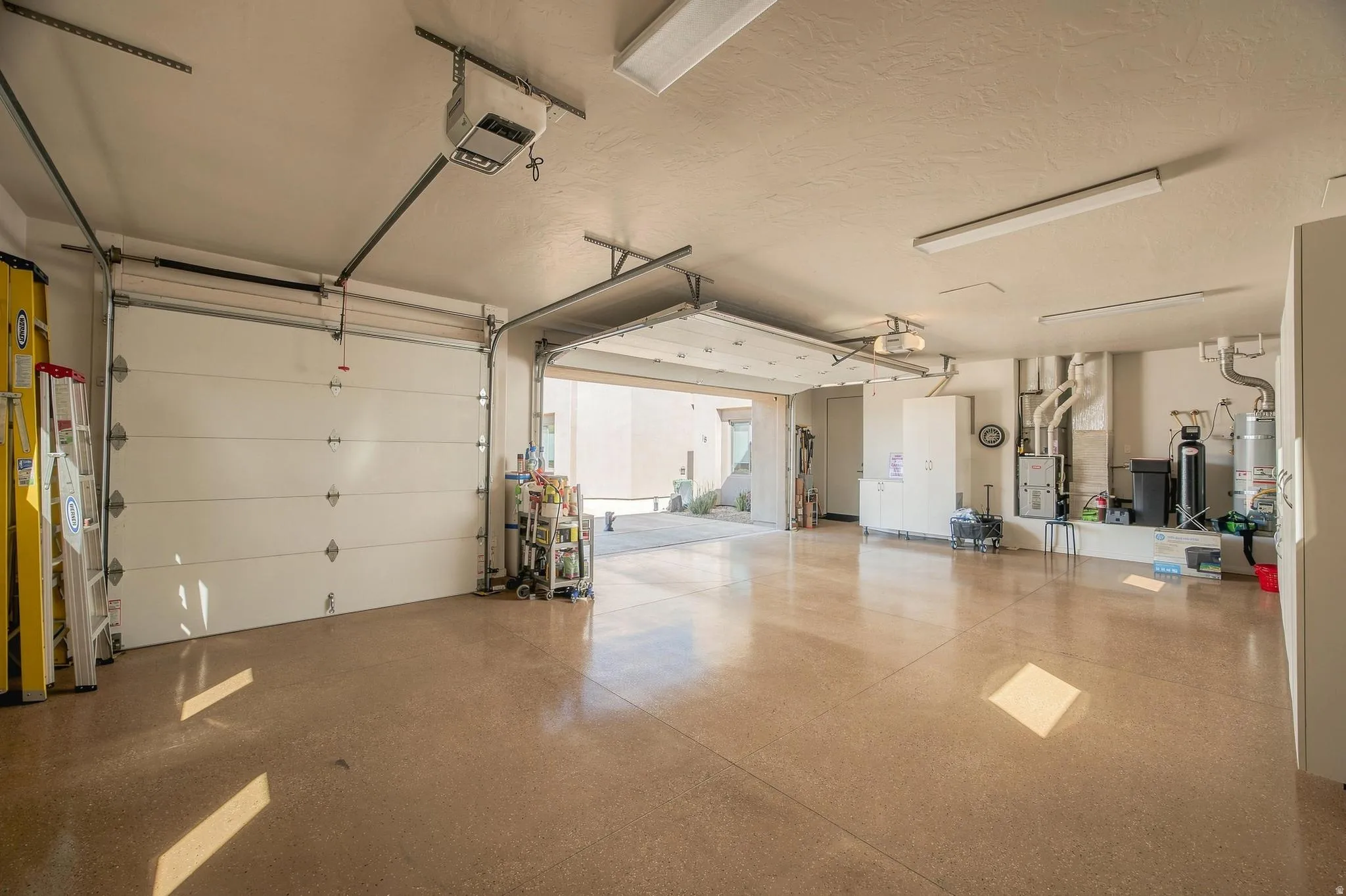 Spacious multi-bay garage with epoxy-coated flooring, multiple overhead fluorescent light fixtures, and a utility area featuring a water heater