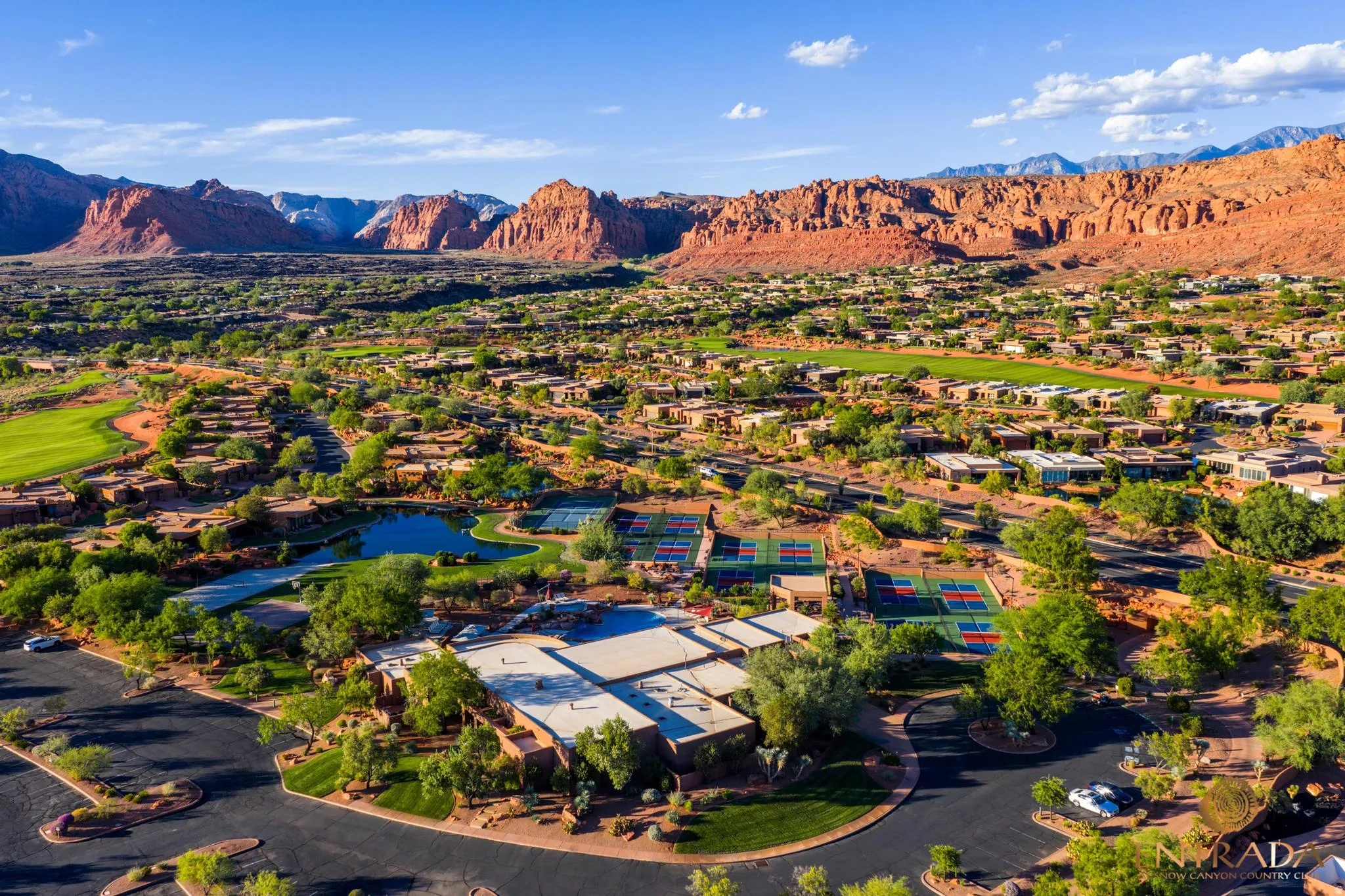 Community featuring multiple tennis courts, a central pond, surrounding residential structures, and a golf course, all set against a prominent red rock mountain backdrop