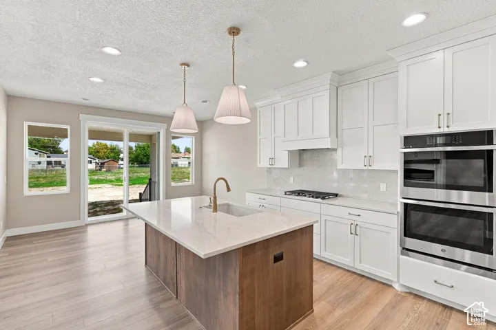 Kitchen featuring white cabinetry, stainless steel double wall ovens, a central island with a sink, pendant lighting, and wood-finish flooring