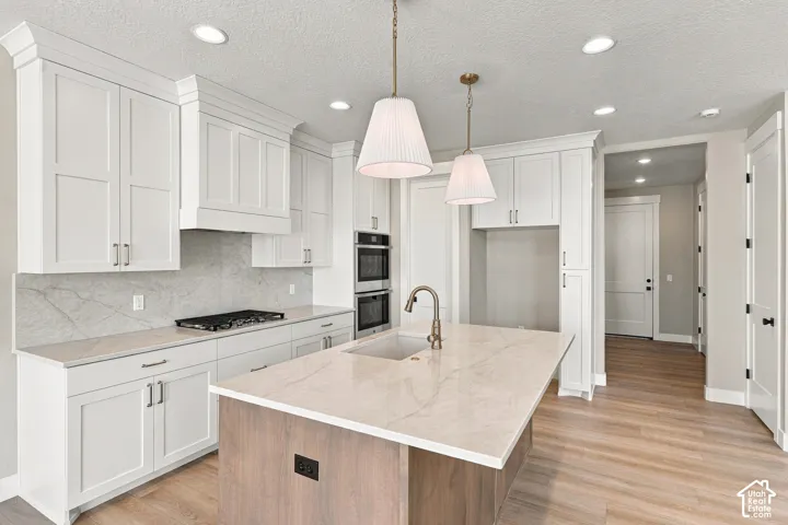 Modern kitchen featuring white shaker cabinetry, light-toned wood-finish flooring, a central island with a sink, double wall ovens, and a gas cooktop