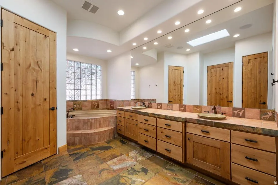 Dual vanity with integrated sinks, recessed lighting, wood-finish cabinetry, a built-in bathtub, and a glass block window