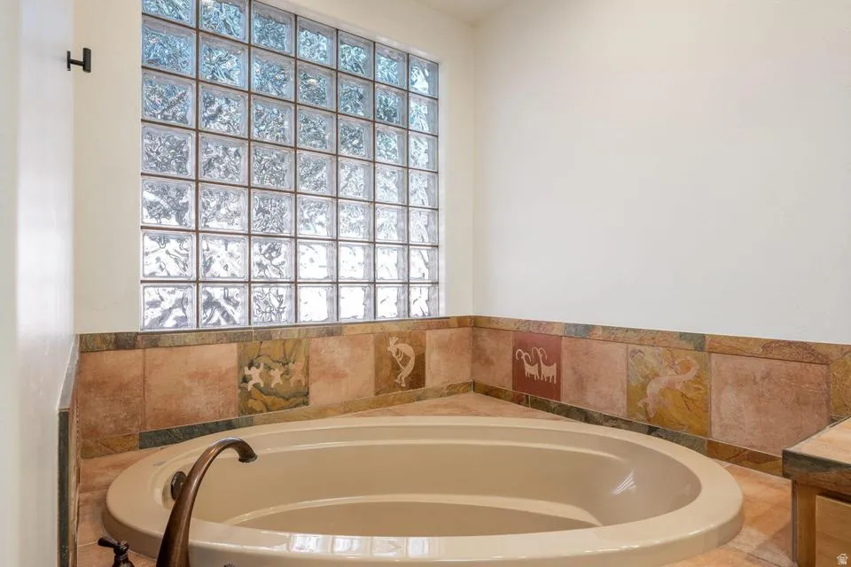 Built-in corner tub with bronze-tone fixtures, surrounded by decorative tile work and a glass block window