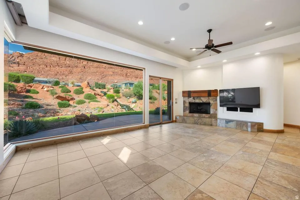 Expansive window wall, stone-clad fireplace with wood mantle, large format tile flooring, recessed lighting, and ceiling fan