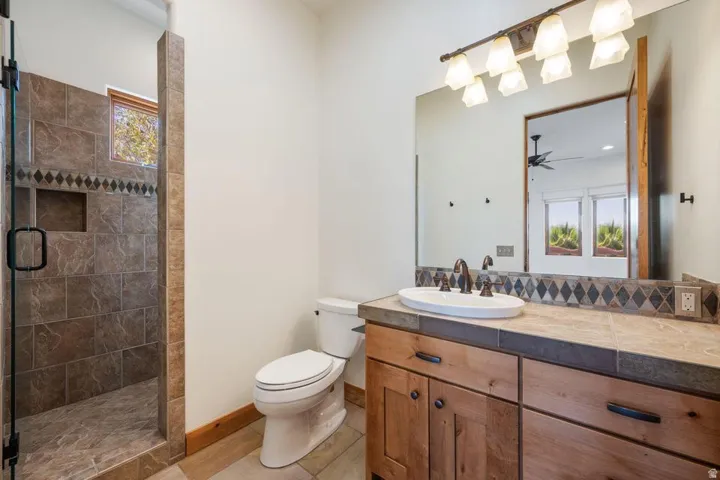 Walk-in shower with tiled surround and built-in niche, wood-finish vanity with an undermount sink, tiled countertop, and a large vanity mirror with overhead lighting