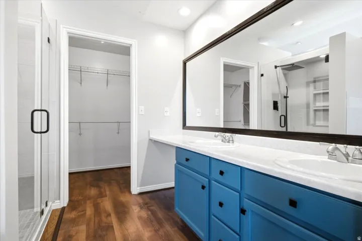 Dual basin vanity with blue cabinetry, a full-width mirror, a glass-enclosed shower, and wood-finish flooring