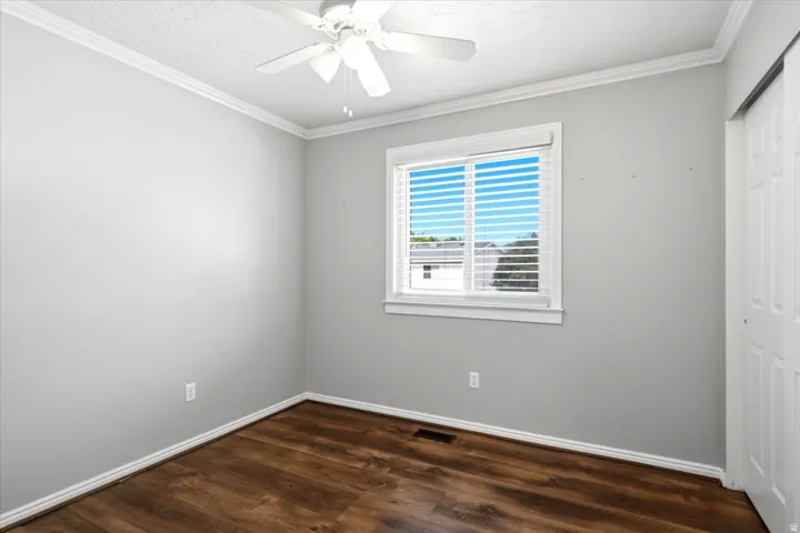 Wood-finish flooring, white ceiling fan, crown molding, double-pane window with blinds, and a sliding closet door